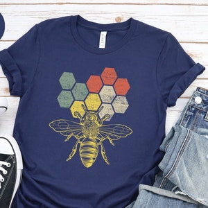 Puede incluir: Una camiseta azul marino con un gráfico de estilo vintage de una abeja y un panal. El panal está formado por seis hexágonos en tonos de verde, amarillo, naranja y rojo.