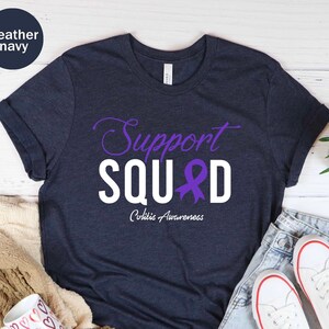 May include: A heather navy blue t-shirt with the text "Support Squad" in white letters with a purple ribbon in the middle of the word "Squad". The text "Colitis Awareness" is written in smaller white letters below the word "Squad".