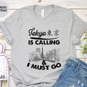 Może przedstawiać: Szary T-shirt z napisem "Tokyo 東京 IS CALLING & I MUST GO" w kolorze czarnym. Na koszulce znajduje się czarno-biała ilustracja miejskiego krajobrazu z wieżą w centrum.