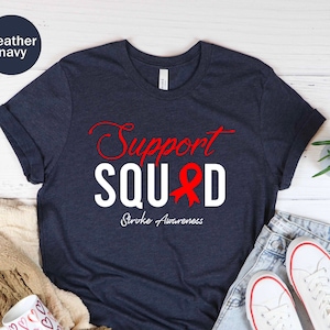 Op de afbeelding: Een heideblauw t-shirt met de tekst "Support Squad" in witte letters en een rood lint. De tekst "Stroke Awareness" staat in kleinere rode letters onder de hoofdtekst.