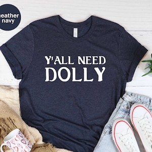 以下が含まれることがあります： ヘザーネイビーブルーのTシャツに、白い文字で「Y'all Need Dolly」とプリントされています。