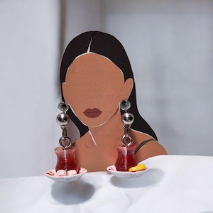 May include: A pair of unique earrings featuring miniature red tea glasses with silver-toned metal accents. Each glass rests on a small white saucer with a delicate design. The earrings hang from a series of silver beads.
