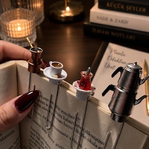 Puede incluir: Marcadores de metal en miniatura con temática de café. El juego incluye una cafetera de cobre, una taza de té blanca con un platillo y un vaso de té con una cuchara. Los marcadores están sujetos a las páginas de un libro.