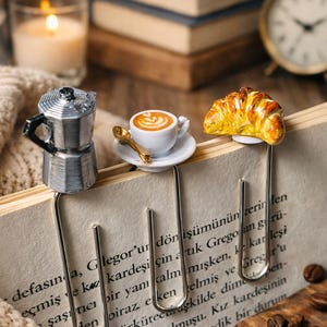 May include: Three metal page markers attached to the top of an open book. The left marker features a silver moka pot, the center marker has a miniature white coffee cup with latte art and a gold spoon, and the right marker displays a croissant.