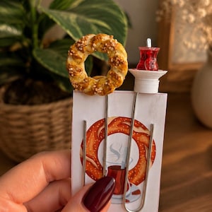 May include: A set of two metal paper clips attached to a rectangular card. One clip features a miniature golden-brown braided bagel with salt crystals, and the other features a small red tea glass on a white saucer. The card displays a graphic illustration.