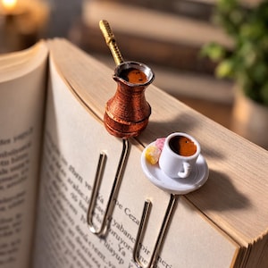 May include: A copper-colored miniature coffee pot and a white teacup bookmark set on an open book. The coffee pot has a gold handle, and the teacup sits on a white saucer with colorful candies. The bookmark is made of metal.