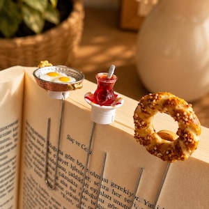 Könnte beinhalten: Drei Metall-Lesezeichen mit Miniatur-Lebensmitteln: eine kleine Pfanne mit zwei Spiegeleiern, ein Glas roter Tee auf einer weißen Untertasse mit einem winzigen Löffel und ein goldbrauner, mit Sesam bestreuter Bagel, befestigt an einem Buch.
