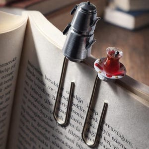May include: A silver-coloured metal bookmark, shaped like a Turkish tea pot and a small glass of tea, on the pages of a book. The tea glass contains a red liquid. The book is open, with text visible on the pages.