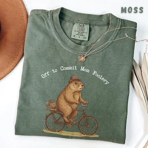 May include: A moss green t-shirt featuring the text "Off to Commit Mom Foolery" and an illustration of a groundhog on a bicycle, wearing a hat and carrying flowers. The shirt has a Comfort Colors tag.