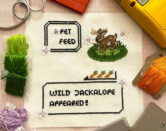 Op de afbeelding: Een kruissteekborduurwerk op witte stof met een gepixelde bruine jackalope op een groen vlak, tekst met PET FEED en WILD JACKALOPE APPEARED!, omringd door groen, oranje en lavendelkleurig borduurgaren, een schaar en spelcartridges.