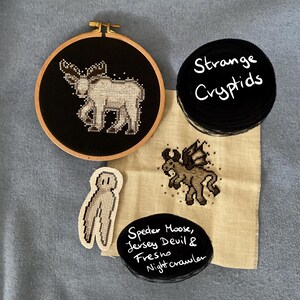 Peut inclure: Trois cerceaux à broder avec un tissu noir et une broderie blanche de cryptides. Les cerceaux sont étiquetés "Strange Cryptids", "Speeder Moose, Jersey Devil & Fresno Nightcrawler", et montrent un élan, une créature ailée et une créature aux longues pattes.