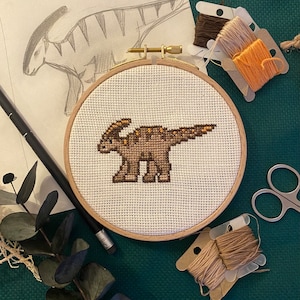 Peut inclure: Un dinosaure brodé de couleur marron et orange sur une toile blanche dans un cercle de broderie en bois. Le cercle est sur une surface verte avec du fil à broder et des ciseaux.