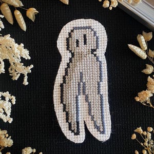 May include: A white cross-stitch embroidery of a grey ghost with a simple face. The ghost is standing on a black background.