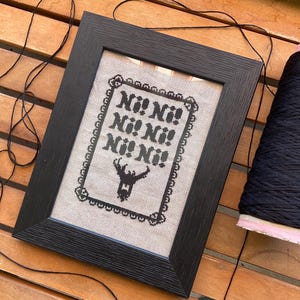 May include: Black framed cross-stitch artwork with the phrase "Ni! Ni! Ni! Ni! Ni!" in black on a beige fabric. A black deer silhouette is below the text. A black thread spool is on the right. The frame is black.