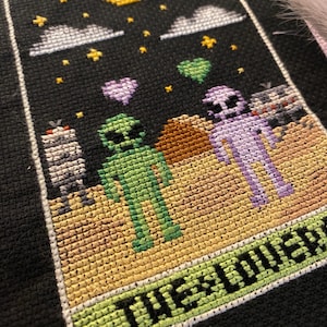 Puede incluir: Arte de punto de cruz que representa la carta del tarot "Los Amantes". El diseño presenta dos figuras alienígenas, una verde y otra morada, con bocadillos en forma de corazón. Una luna creciente, estrellas y nubes en el cielo negro. El texto "THE LOVERS" está en la parte inferior.