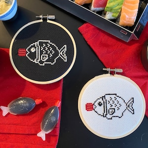 May include: Two cross-stitch hoops with a fish design, one on black fabric and one on white. The fish is outlined in white thread with a red mouth. Soy sauce packets and sushi are also visible.