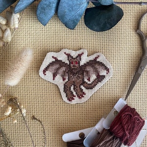 May include: A cross-stitch embroidery of a brown bat with red eyes. The bat is stitched on a beige Aida cloth. The embroidery is surrounded by brown and red embroidery floss.