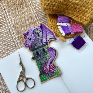 Peut inclure: Un motif de dragon en point de croix violet, gris, vert et rose, avec une bordure beige. Le dragon est sur une tour verte. De petits ciseaux et des bobines de fil à broder violets et roses sont également visibles.