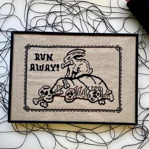 May include: Framed cross-stitch art with a black border. The design features a rabbit holding a bone, atop a pile of skulls and bones. The text "RUN AWAY!" is above the rabbit in black letters. The background is a light tan color.