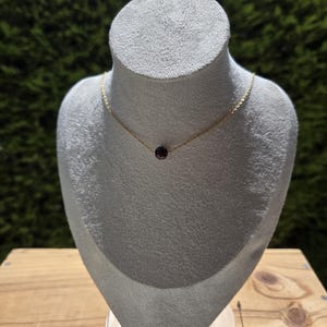 Peut inclure: Un collier à chaîne fine en or avec une perle unique, petite et ronde, de couleur bordeaux foncé au centre. Le collier est présenté sur un présentoir à bijoux texturé de couleur blanc cassé, mettant en valeur le contraste entre le métal doré et la pierre rouge.