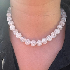 May include: A clear, beaded necklace with a crackled texture. The necklace is composed of round, translucent beads, strung together to form a choker style. The necklace is worn against a dark background.
