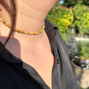 Op de afbeelding: Een choker ketting met kleine, ronde, gele en bruine kralen. De ketting wordt gedragen over een zwart overhemd, met een vage groene achtergrond van bladeren en zonlicht.