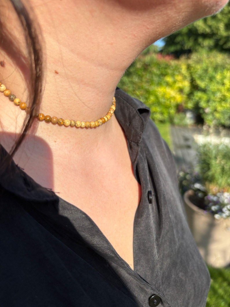 Op de afbeelding: Een choker ketting met kleine, ronde, gele en bruine kralen. De ketting wordt gedragen over een zwart overhemd, met een vage groene achtergrond van bladeren en zonlicht.