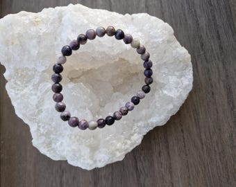 Könnte beinhalten: Ein violettes und weißes Perlenarmband auf einem weißen Kristallstein. Das Armband besteht aus kleinen, runden Perlen.