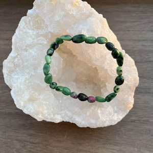 Può includere: Un braccialetto di pietre preziose verdi e rosa con perline di forma ovale. Il braccialetto è appoggiato su un cristallo bianco.