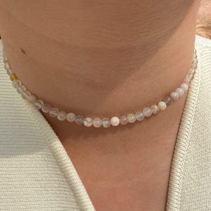 May include: A delicate beaded necklace composed of small, round, light-colored beads. The necklace is worn around the neck, resting against a cream-colored top. The beads appear to be a mix of pale pink, white, and beige.