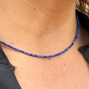 May include: A delicate necklace featuring small, round, deep blue beads. The necklace is worn close to the neck, creating a simple and elegant look. The beads appear to be evenly sized and strung together, forming a continuous strand. Jewelry.