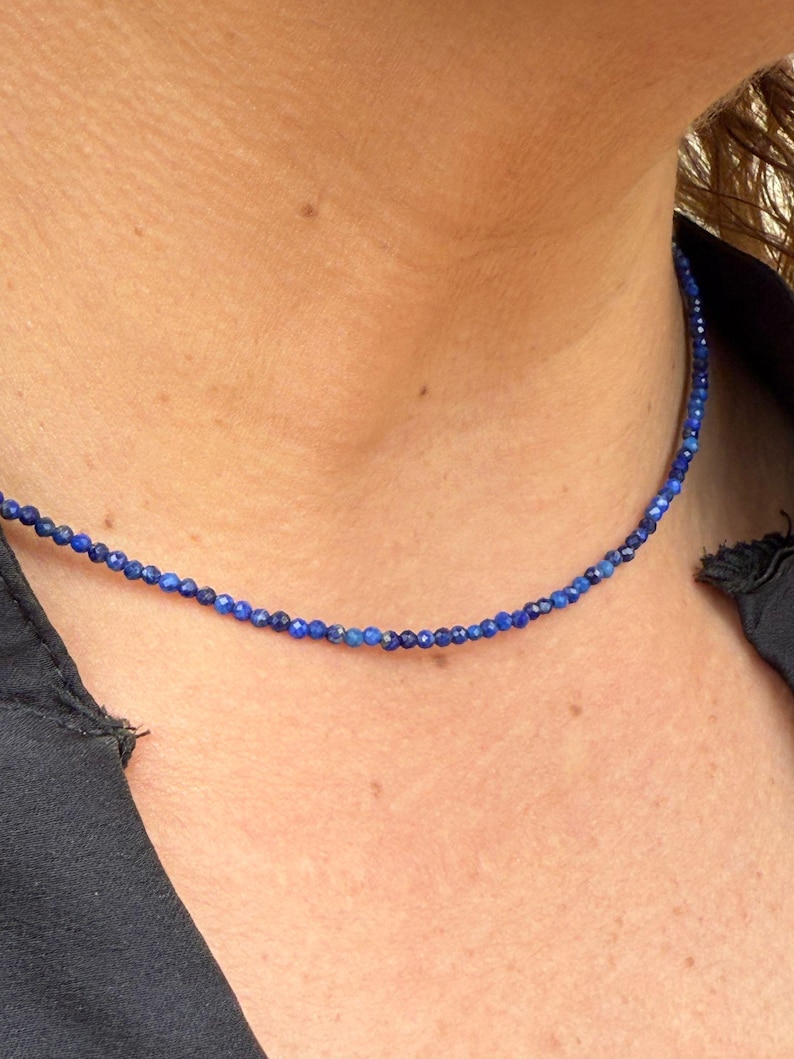 May include: A delicate necklace featuring small, round, deep blue beads. The necklace is worn close to the neck, creating a simple and elegant look. The beads appear to be evenly sized and strung together, forming a continuous strand. Jewelry.