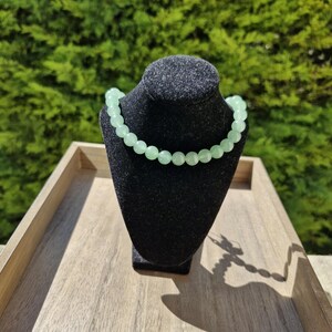 May include: A light green beaded necklace on a black velvet display stand. The necklace is made of round beads and is displayed on a wooden surface.