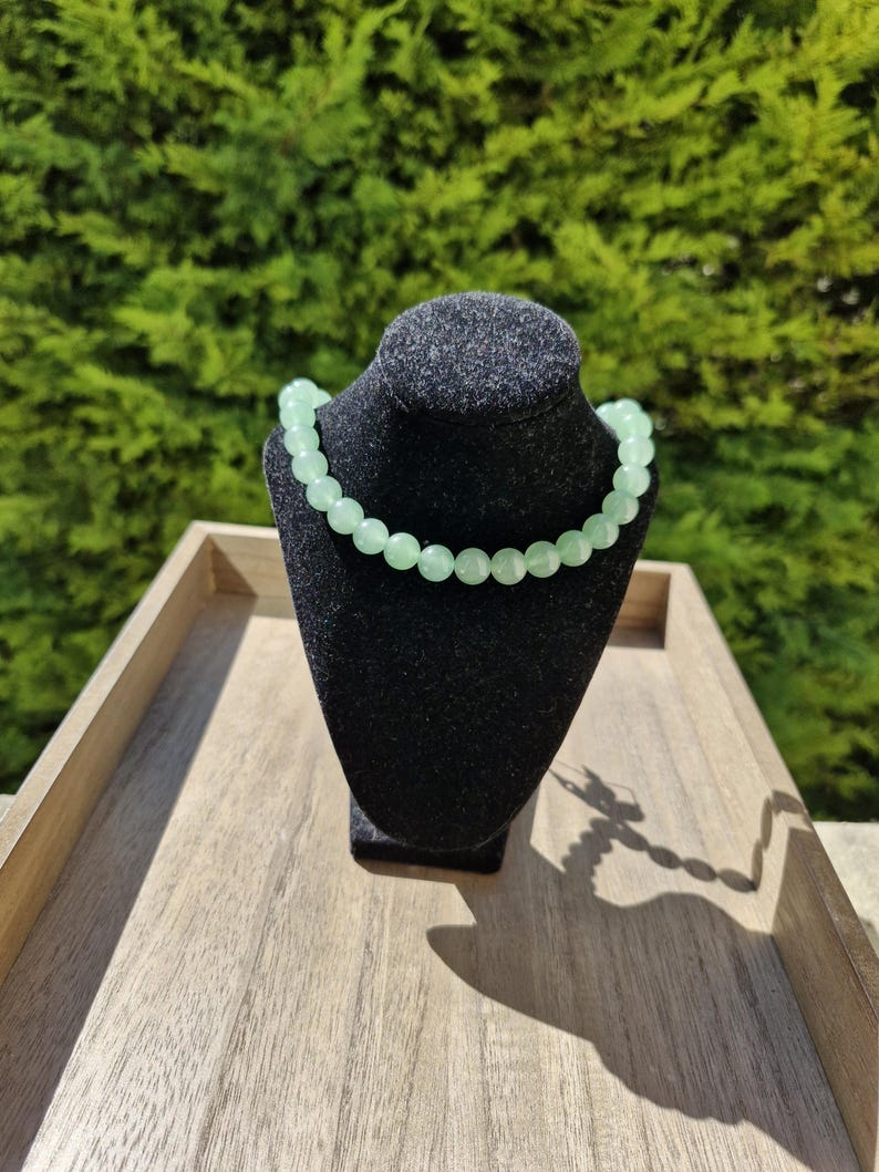 May include: A light green beaded necklace on a black velvet display stand. The necklace is made of round beads and is displayed on a wooden surface.