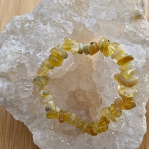 Peut inclure: Un bracelet composé de petites pierres précieuses jaunes de forme irrégulière. Le bracelet est posé sur une surface blanche texturée, avec un fond en bois. Les pierres présentent différentes nuances de jaune.