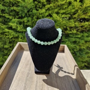 May include: A light green beaded necklace on a black display stand. The necklace is made of round beads and is on a wooden surface.