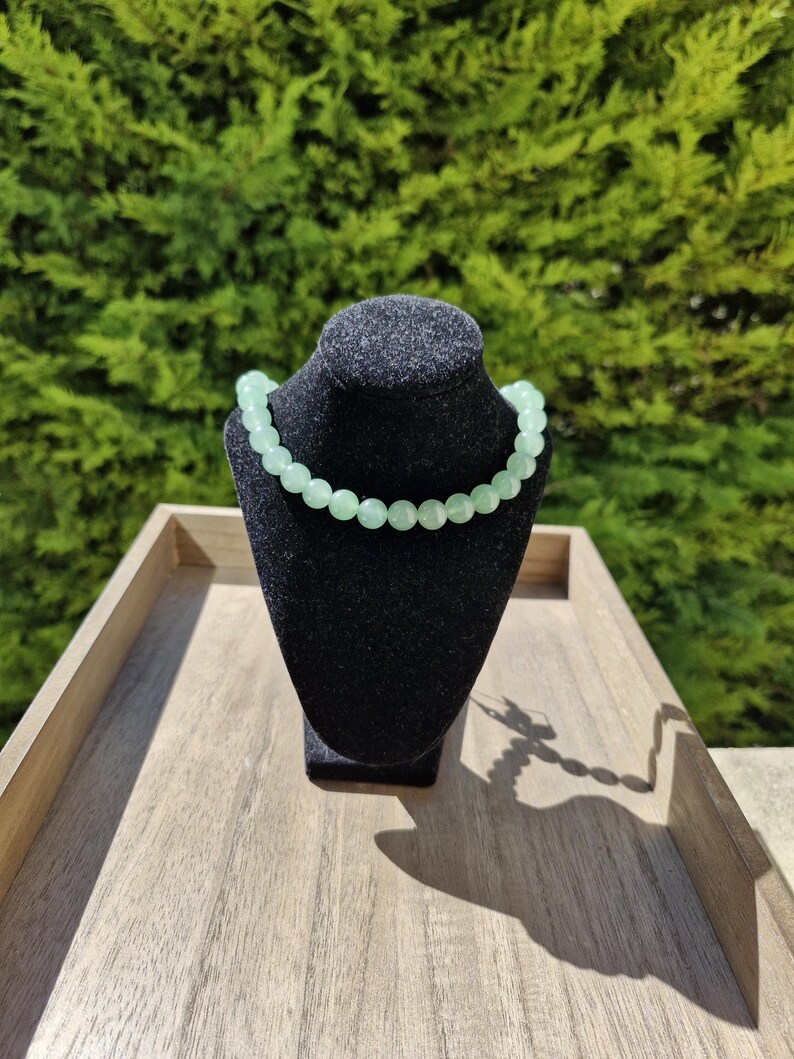 May include: A light green beaded necklace on a black display stand. The necklace is made of round beads and is on a wooden surface.
