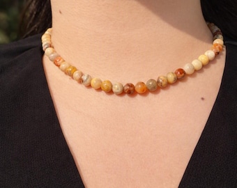 May include: A beaded choker necklace featuring small, round, polished stones in a variegated color palette of warm amber, cream, tan, and reddish-brown tones. The beads are uniform in size and strung together to form a continuous circular band.
