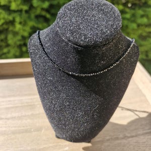 May include: A fine black beaded necklace displayed on a black bust. The necklace features small, dark beads, forming a subtle, elegant design. The bust is set against a blurred green backdrop.