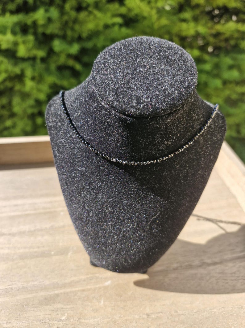 May include: A fine black beaded necklace displayed on a black bust. The necklace features small, dark beads, forming a subtle, elegant design. The bust is set against a blurred green backdrop.