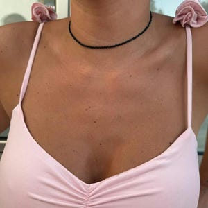 May include: A black beaded choker necklace is worn against a light pink top. The top features spaghetti straps with fabric flower details and a ruched design. The image is a close-up.