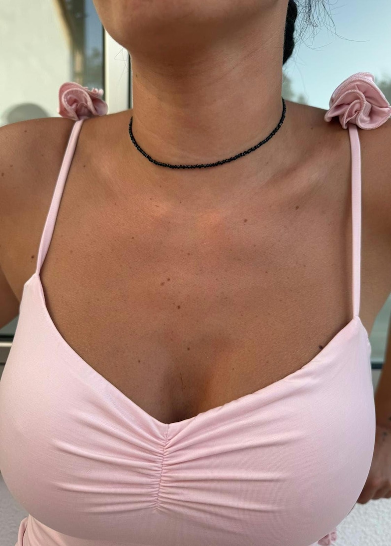 May include: A black beaded choker necklace is worn against a light pink top. The top features spaghetti straps with fabric flower details and a ruched design. The image is a close-up.