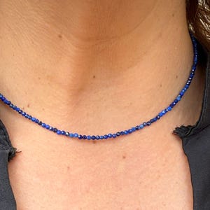May include: A delicate necklace composed of small, deep blue beads. The beads are strung together to form a choker-style necklace. The necklace is worn against a dark background, highlighting the vibrant color of the beads. The necklace appears to be made of gemstones.