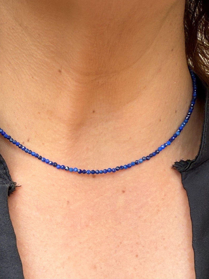 May include: A delicate necklace composed of small, deep blue beads. The beads are strung together to form a choker-style necklace. The necklace is worn against a dark background, highlighting the vibrant color of the beads. The necklace appears to be made of gemstones.