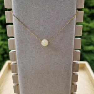 Op de afbeelding: Een delicate gouden ketting met een enkele, ronde, cr&egrave;mekleurige parel hanger. De ketting wordt getoond op een grijze fluwelen sieradenstandaard met een houten frame, tegen een vage groene achtergrond.