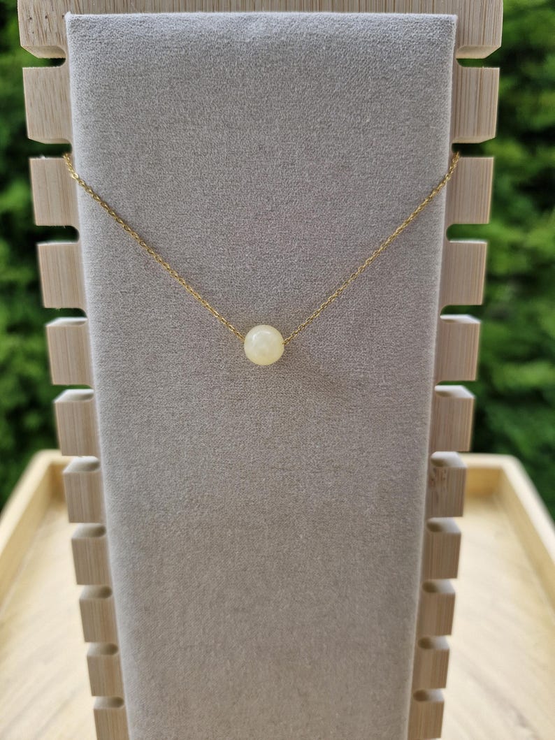 Op de afbeelding: Een delicate gouden ketting met een enkele, ronde, cr&egrave;mekleurige parel hanger. De ketting wordt getoond op een grijze fluwelen sieradenstandaard met een houten frame, tegen een vage groene achtergrond.