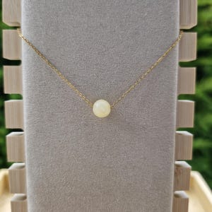 Op de afbeelding: Een delicate goudkleurige ketting met een enkele, ronde, cr&egrave;mekleurige kralen hanger. De ketting wordt getoond op een grijze fluwelen ondergrond, met een houten displaystandaard op de achtergrond. De ketting is fijn en de kraal is gecentreerd.