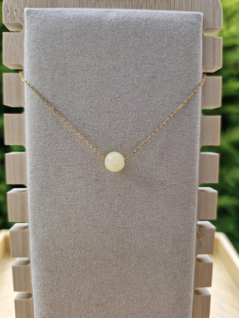 Op de afbeelding: Een delicate goudkleurige ketting met een enkele, ronde, cr&egrave;mekleurige kralen hanger. De ketting wordt getoond op een grijze fluwelen ondergrond, met een houten displaystandaard op de achtergrond. De ketting is fijn en de kraal is gecentreerd.