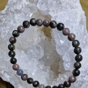 Könnte beinhalten: Ein Armband aus dunkelgrünen und braunen Edelsteinperlen. Die Perlen sind rund und haben ein gesprenkeltes Muster. Das Armband liegt auf einem weißen Kristall.