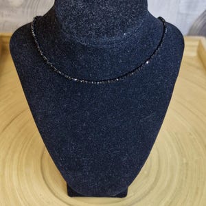 May include: A delicate black beaded necklace displayed on a black velvet jewelry bust. The necklace is composed of small, evenly spaced beads, creating a simple yet elegant design. The bust sits on a light brown wooden surface.
